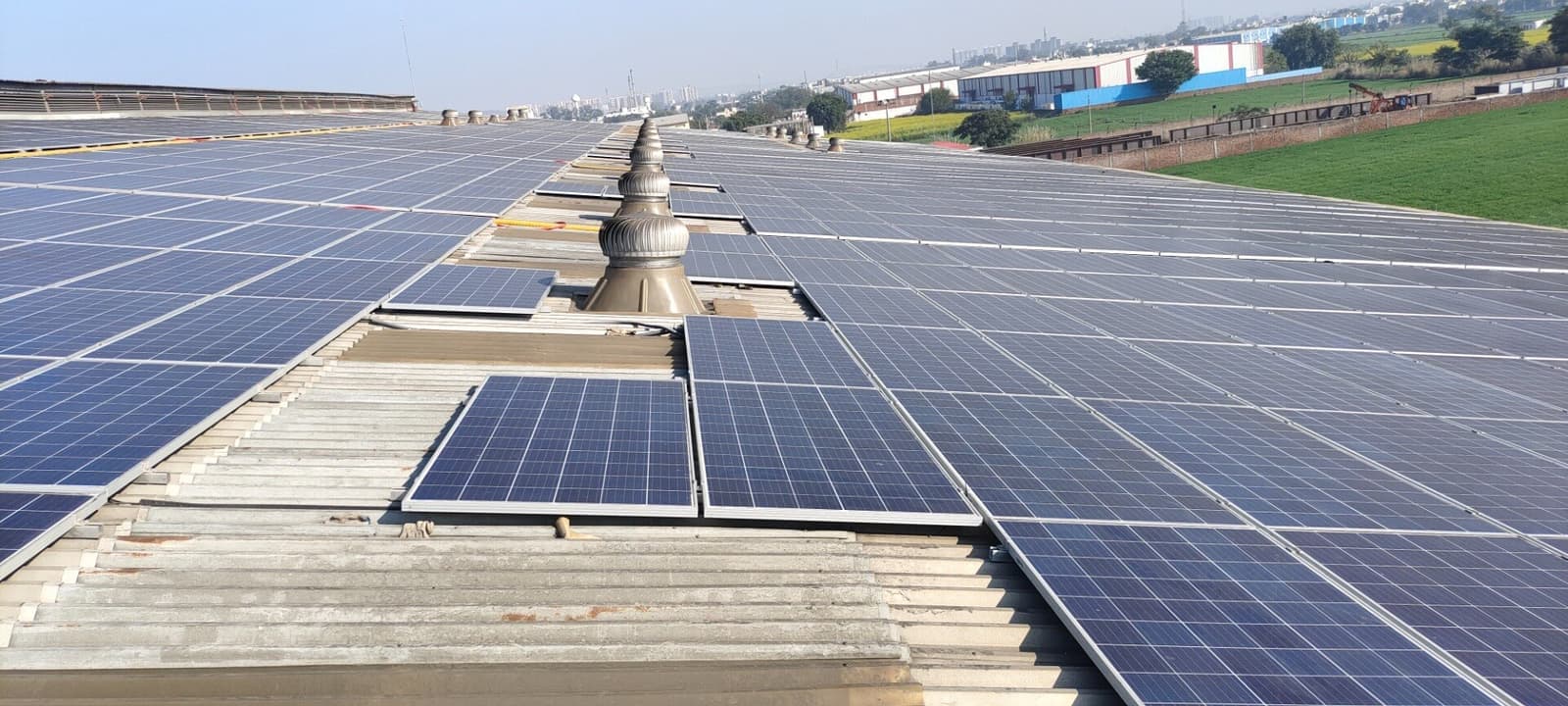 Solar panels covering a long metal-sheet factory roof with turbine ventilators, industrial sheds and green fields beyond.
