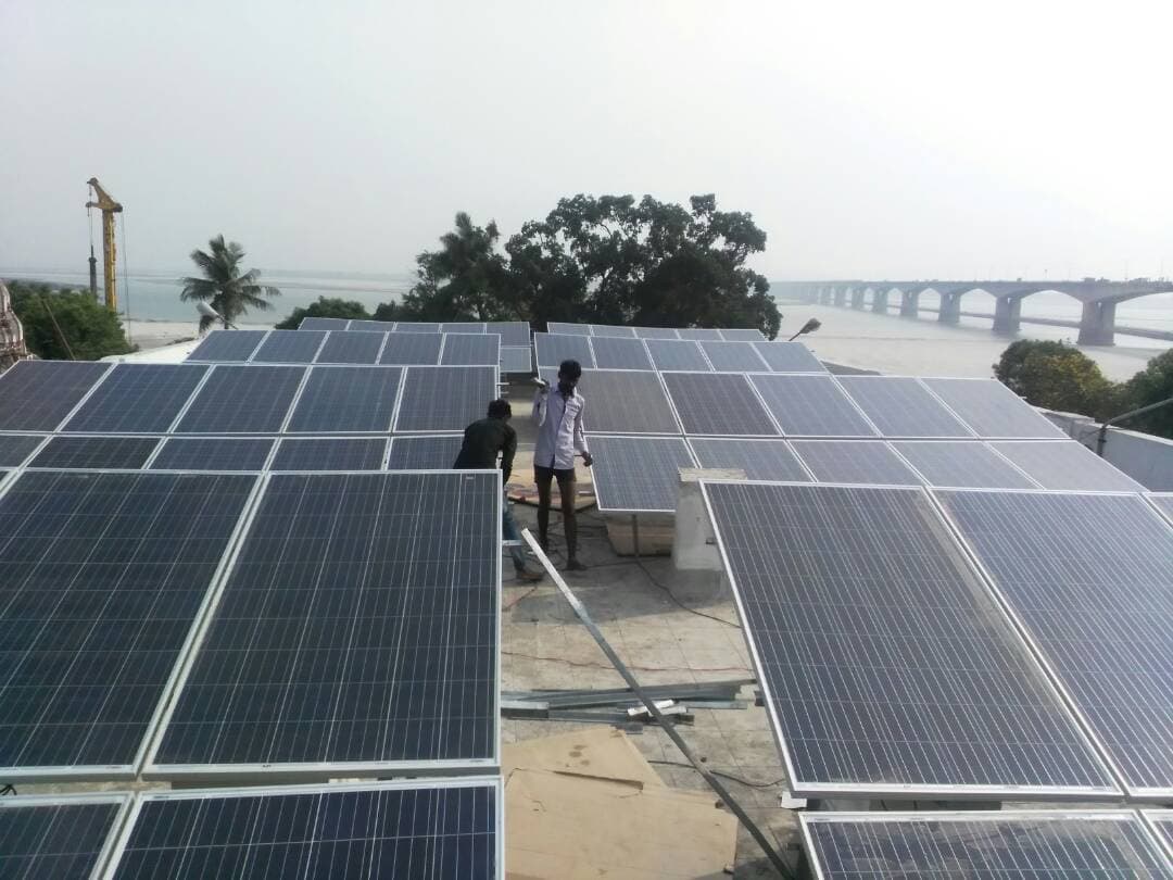 Two workers positioning solar panels on a flat rooftop with a river, a long bridge and a crane behind.