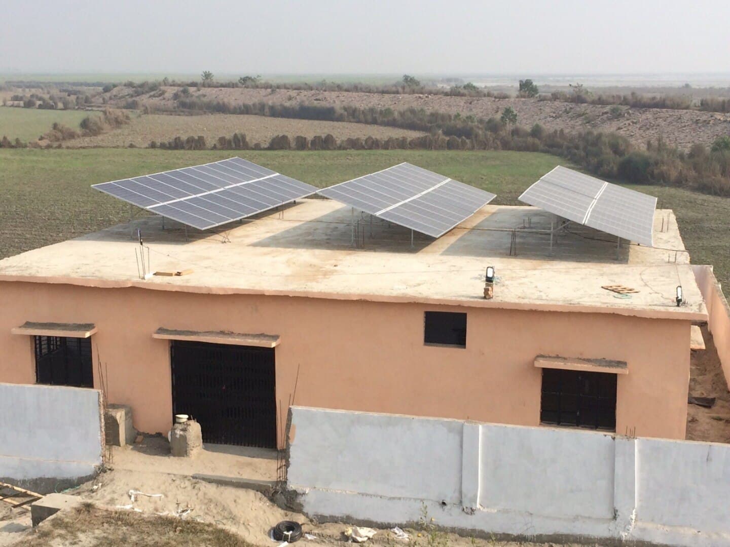 Three tilted solar panel arrays on the flat roof of a small single-storey building in open farmland.