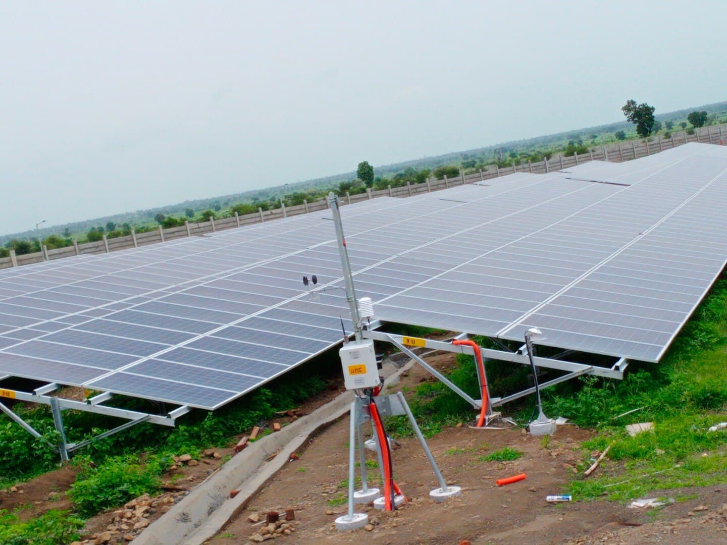 Ground-mounted solar array behind a weather monitoring mast with anemometer and sensors, inside a walled compound.