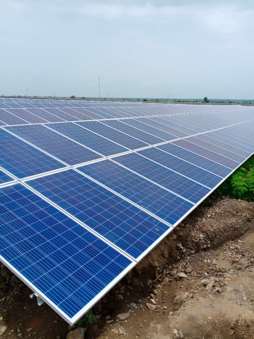 Close view along a row of ground-mounted polycrystalline solar panels on bare earth under an overcast sky.