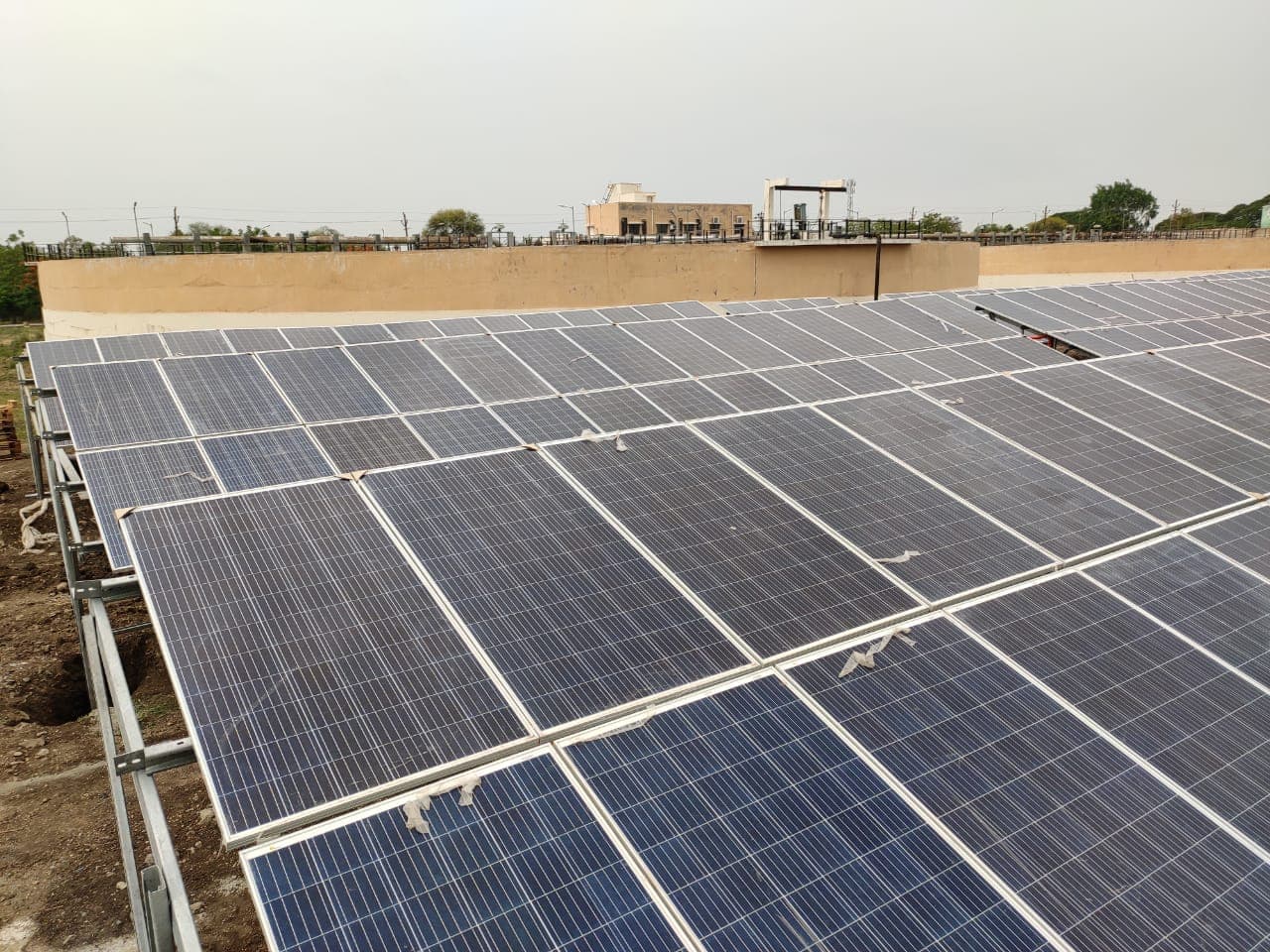 Rows of ground-mounted solar panels on steel rails beside a compound wall, with scraps of tape on the glass.