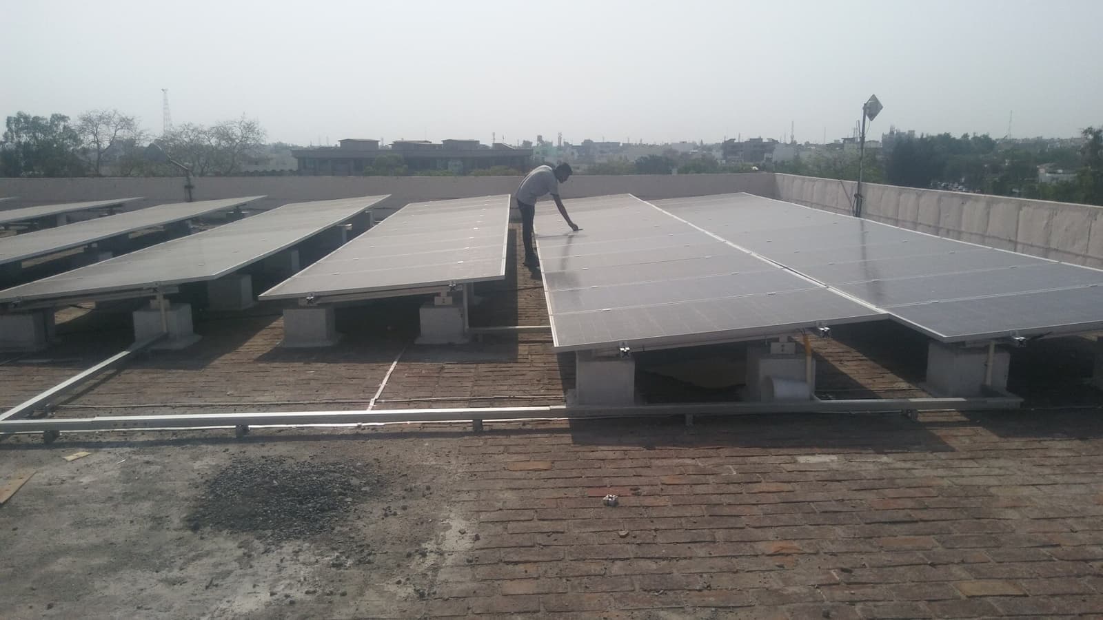 Worker leaning over a solar panel array on concrete plinths on a brick-paved rooftop overlooking a hazy town.