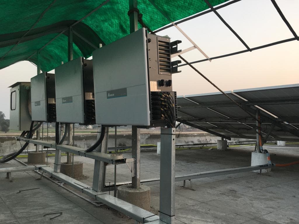 Three string inverters on a steel frame under a green shade net, with an elevated solar array behind.