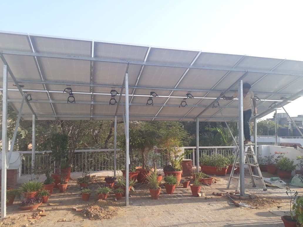Elevated solar panel canopy on galvanised steel posts over a rooftop garden, with an installer on a ladder.