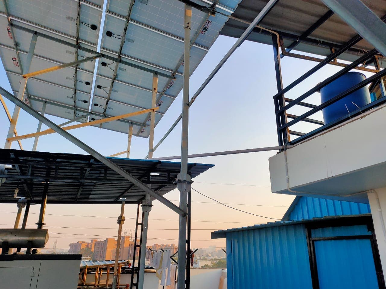 Underside of solar panels on a tall galvanised frame above a rooftop at dusk, water tank behind.