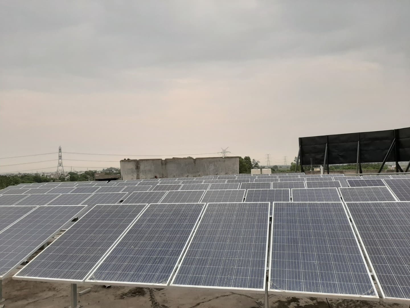 Rooftop solar array with many rows of tilted panels under an overcast sky, transmission towers on the horizon.