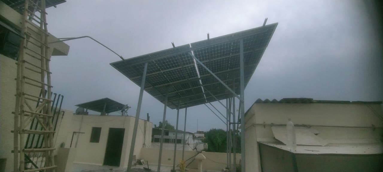 Elevated solar panels on tall steel posts above a house rooftop under an overcast sky, seen from below.