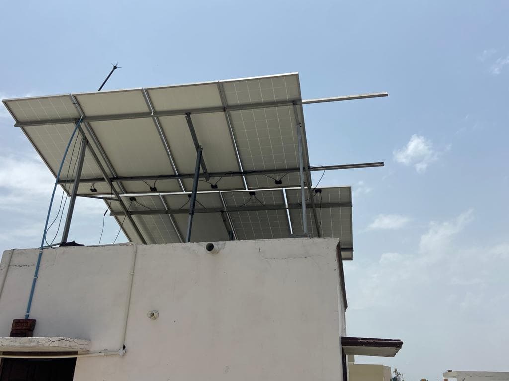 Underside of a raised rooftop solar array on steel posts above a white parapet wall against a clear blue sky.