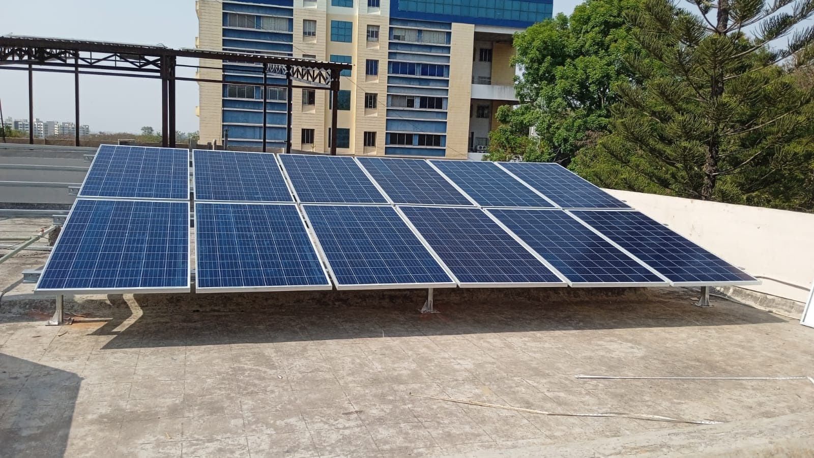 Two rows of blue solar panels on a low steel frame on a flat rooftop beside an office building