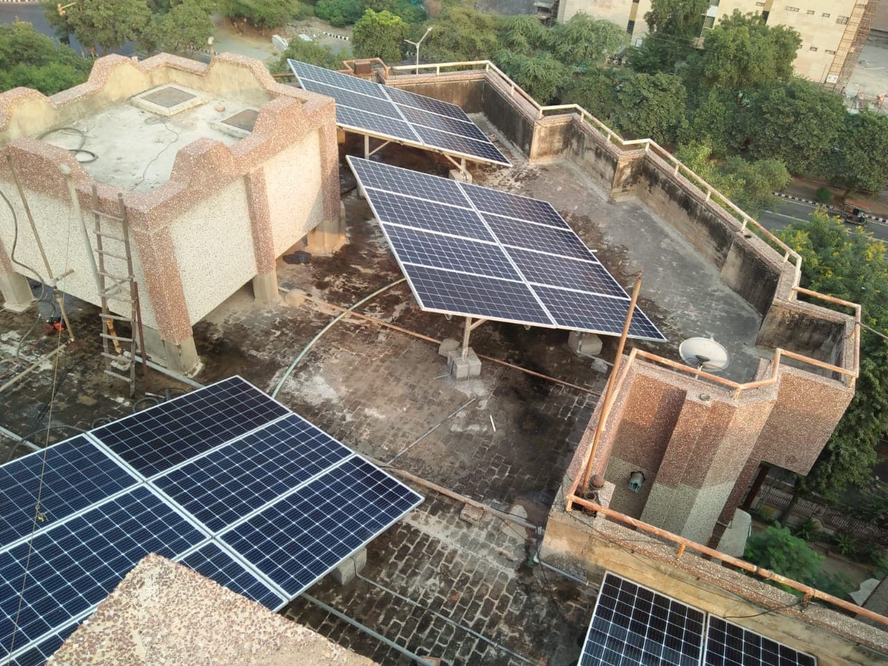 Elevated solar panel arrays on raised steel stands across a flat rooftop, seen from above with trees beyond