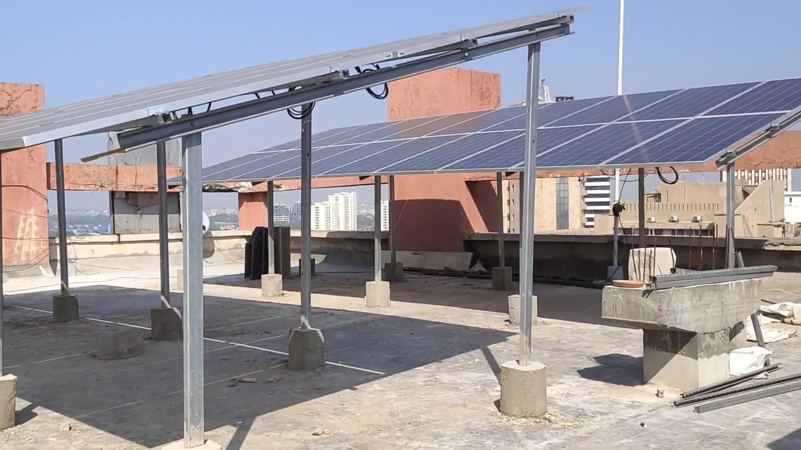 Solar panels on an elevated galvanised steel canopy with concrete footings across a flat rooftop, city buildings behind