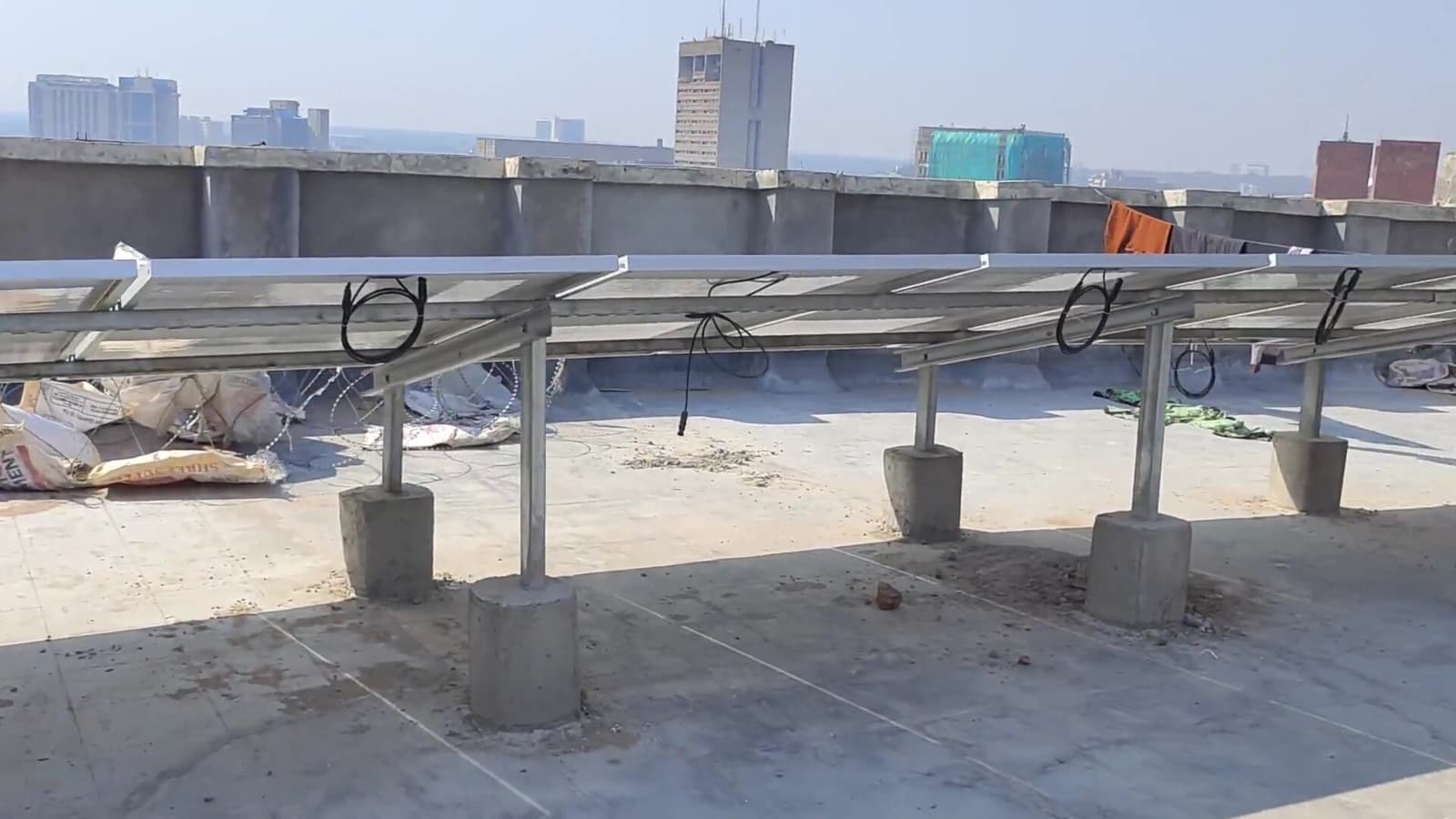 Underside of a solar mounting structure on galvanised posts with concrete footings on a rooftop, with high-rise buildings behind
