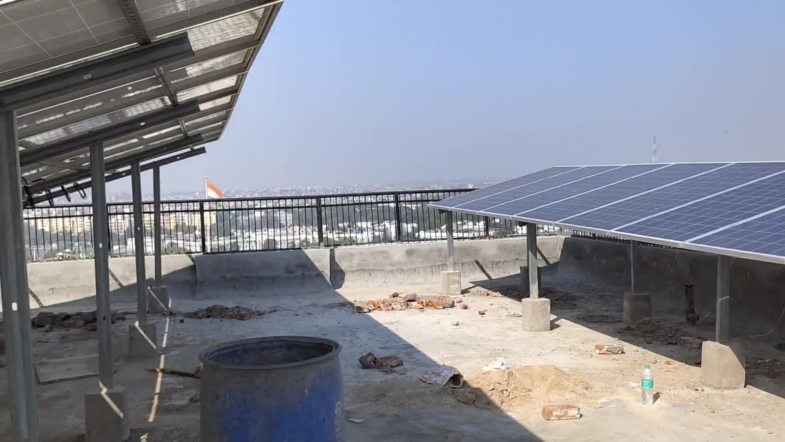 Two elevated solar panel arrays on steel posts over a rooftop under construction, with a city panorama beyond the railing