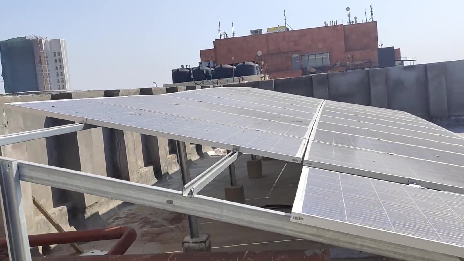 Solar panels on an elevated steel frame at a rooftop edge, with black water tanks and a brick building behind