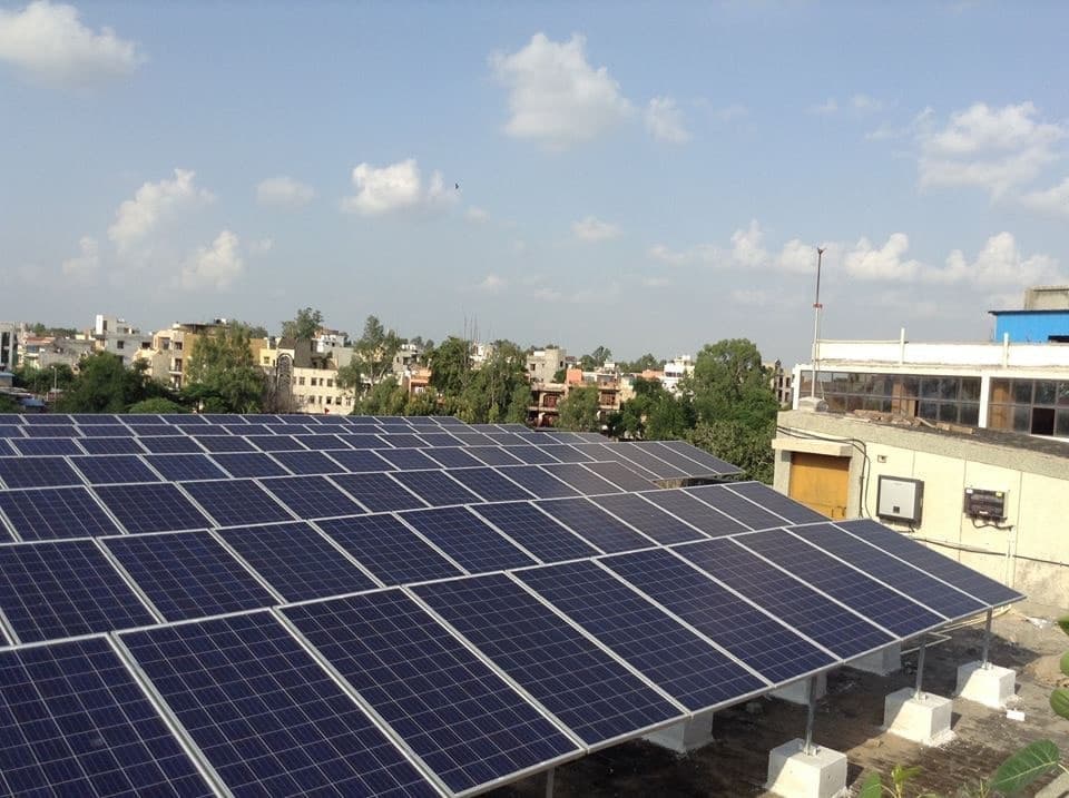 Large rooftop solar array on raised mounts with wall-mounted inverters and a treeline beyond