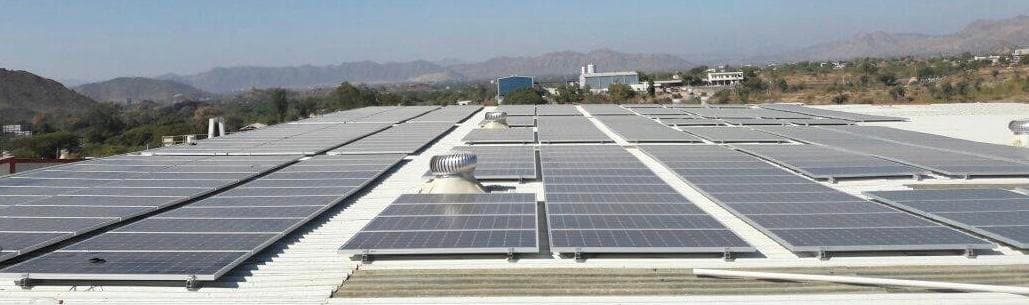 Extensive solar array across a white industrial metal roof with turbine ventilators and hills beyond