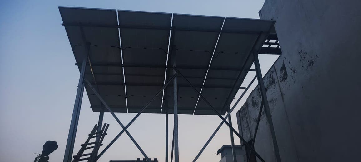 Underside of solar panels mounted on a tall galvanised steel frame beside a plastered wall at dusk