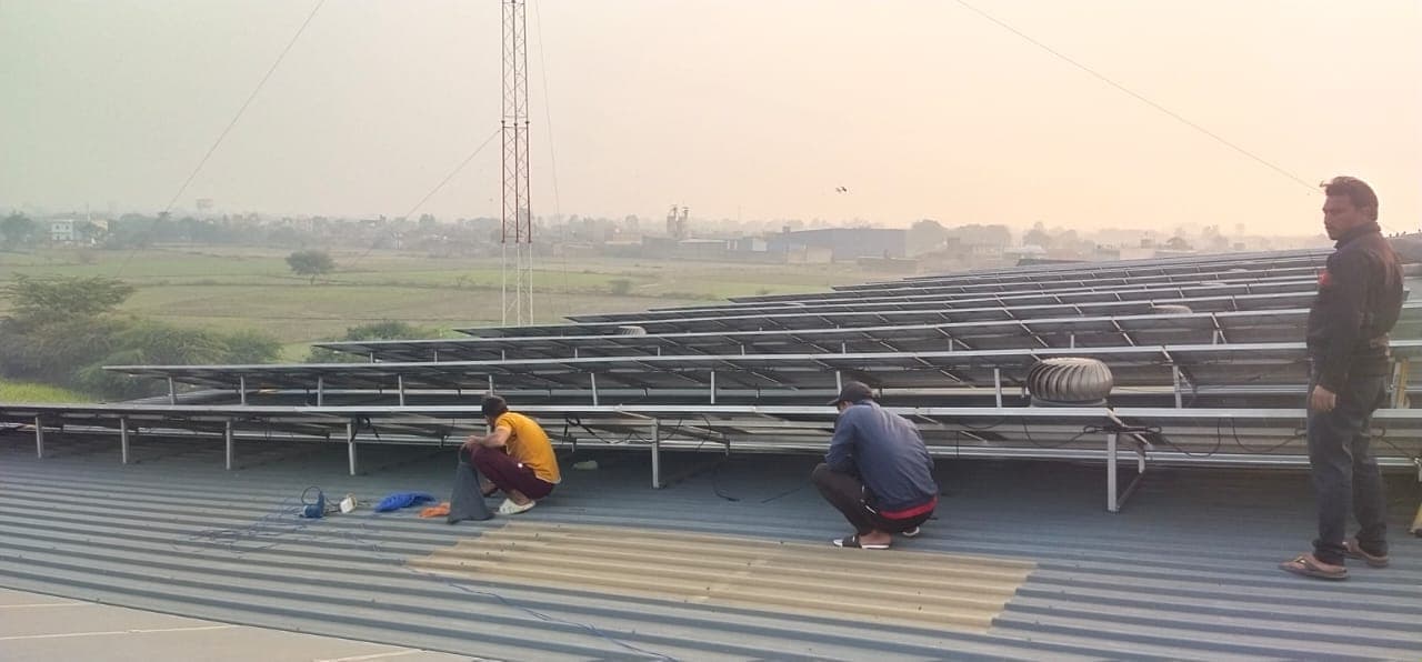Three workers on a corrugated metal roof beside rows of solar panels, with fields in the hazy distance