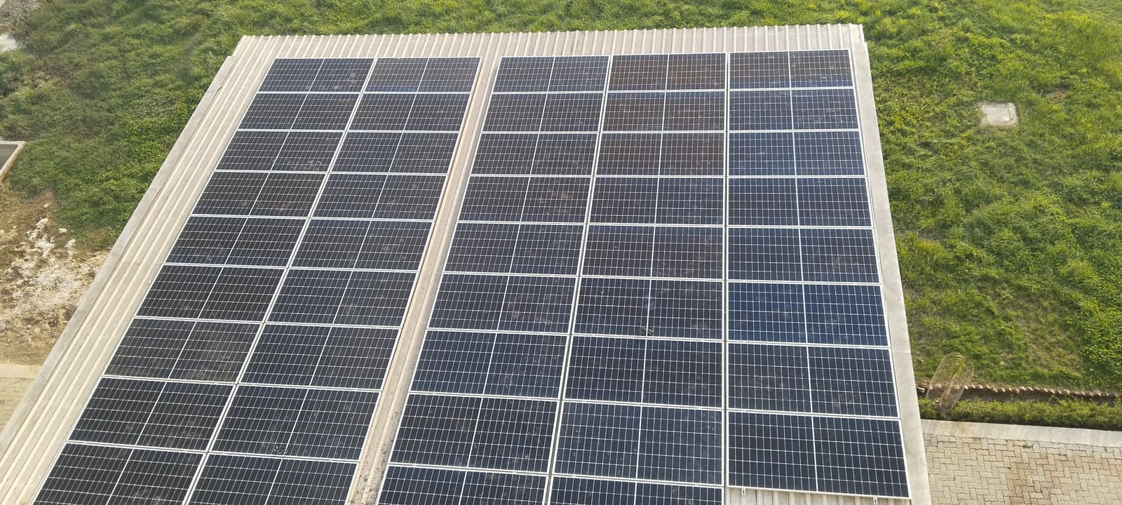 Overhead view of a rectangular block of solar panels on a corrugated roof surrounded by green grass