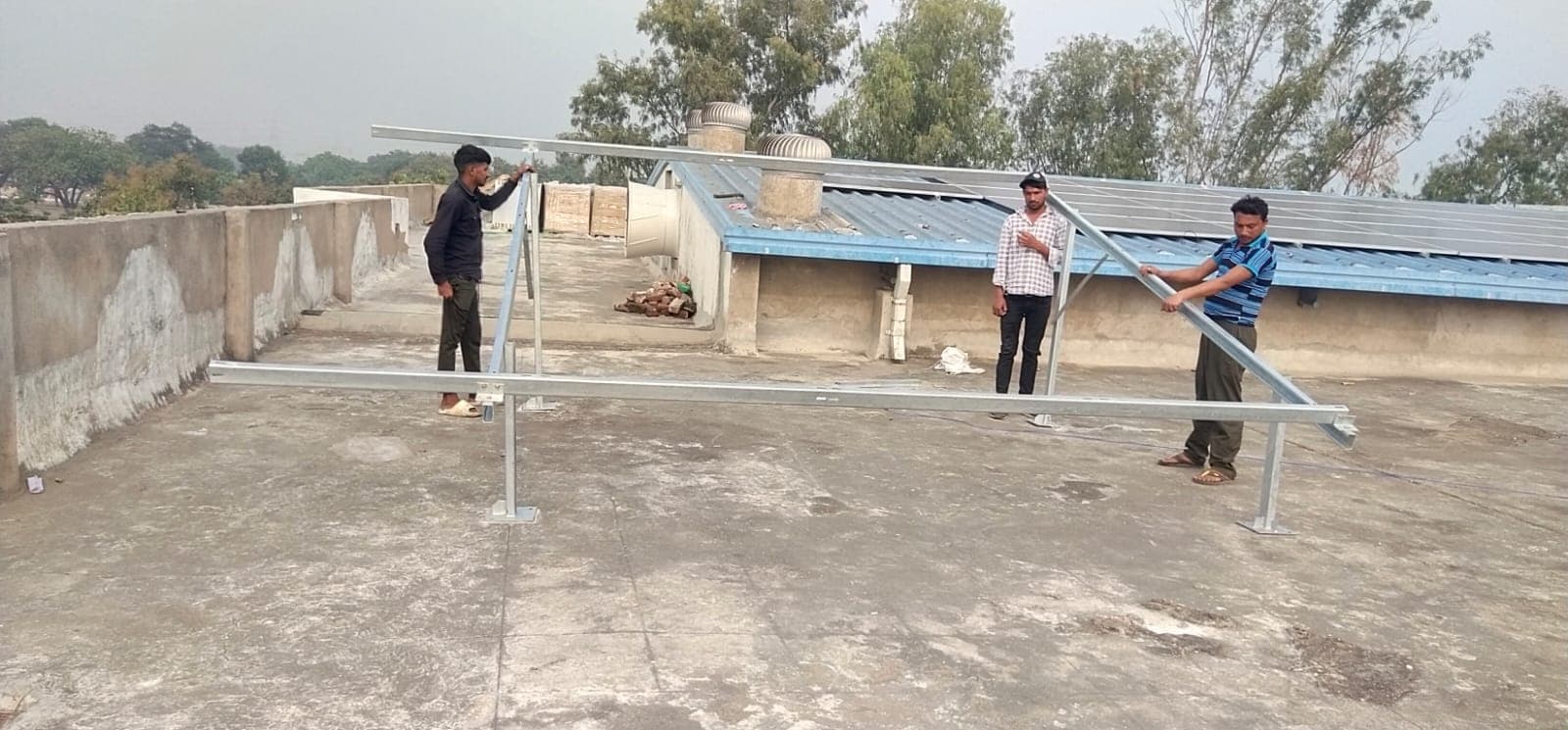 Three workers assembling a galvanised steel mounting frame on a flat concrete rooftop beside a blue-roofed shed