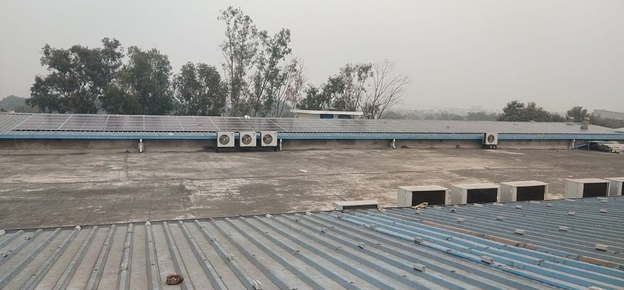 Row of solar panels along a roof edge with air conditioner units below, seen across a flat concrete roof