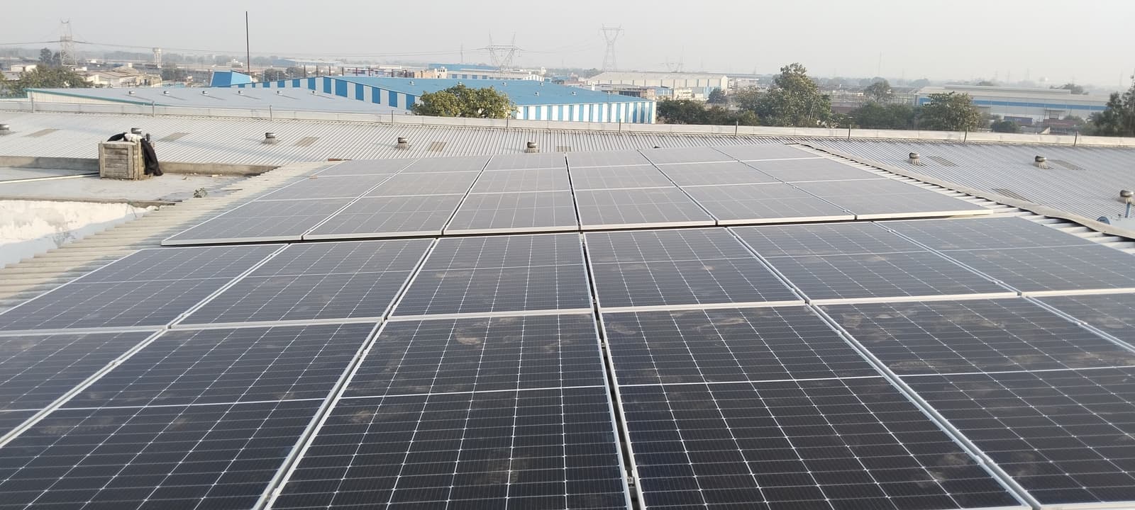 Rows of solar panels on a corrugated metal factory roof with blue-and-white sheds behind under a hazy sky