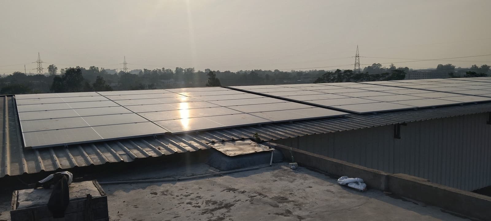 Solar panels on a corrugated roof reflecting the low sun, with trees and transmission towers on the horizon