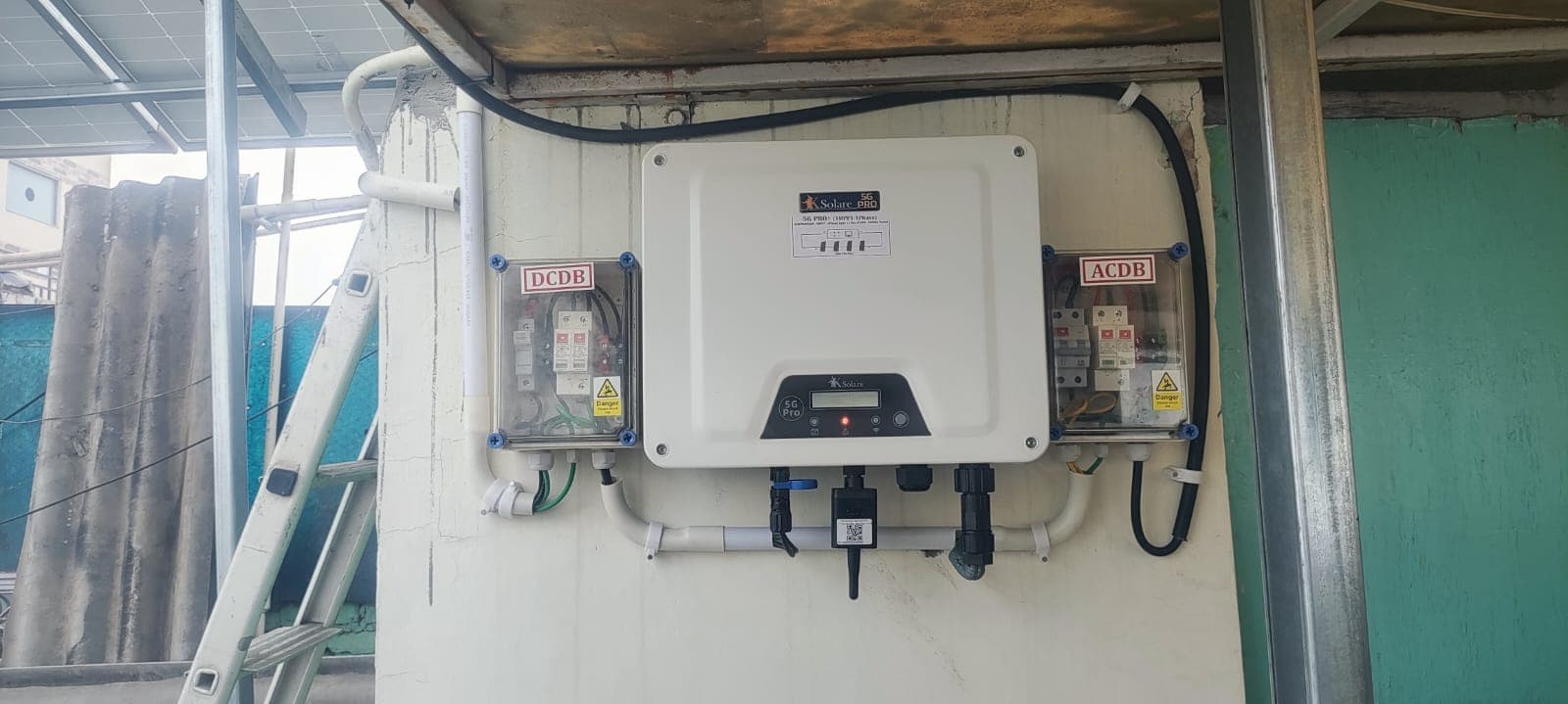 White wall-mounted solar inverter labelled K Solare between DCDB and ACDB junction boxes beside a ladder