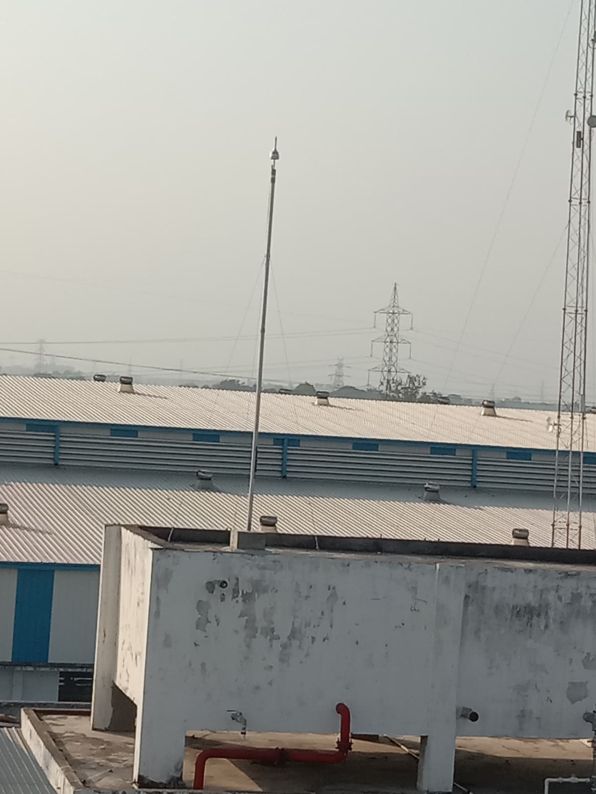 Tall metal mast with guy wires rising above white corrugated factory roofs under a hazy sky