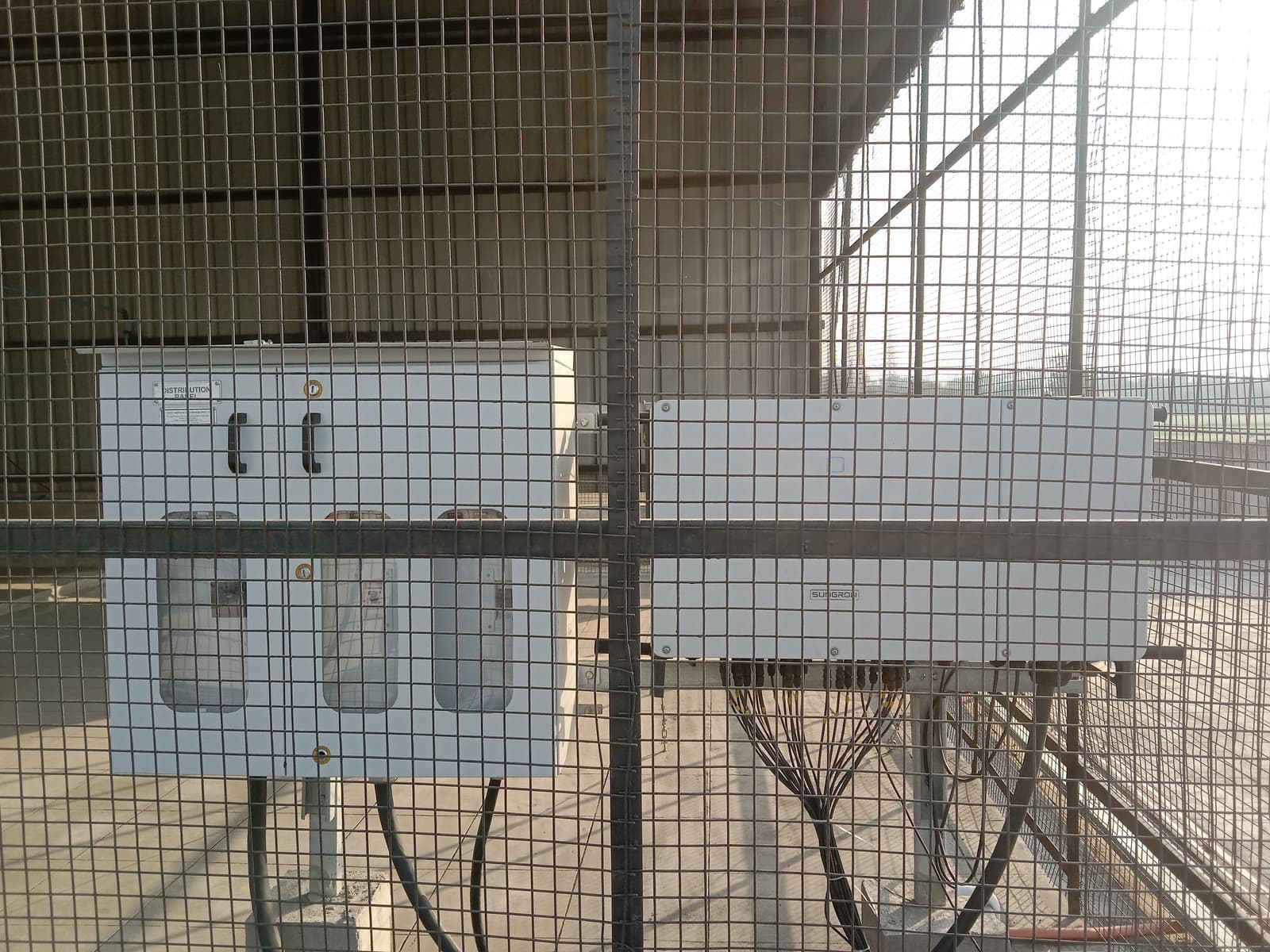 Sungrow inverter and distribution panel mounted inside a wire mesh enclosure under a factory roof