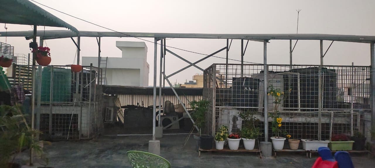 Elevated galvanised steel mounting structure over a rooftop terrace with caged water tanks, potted plants and hazy sky