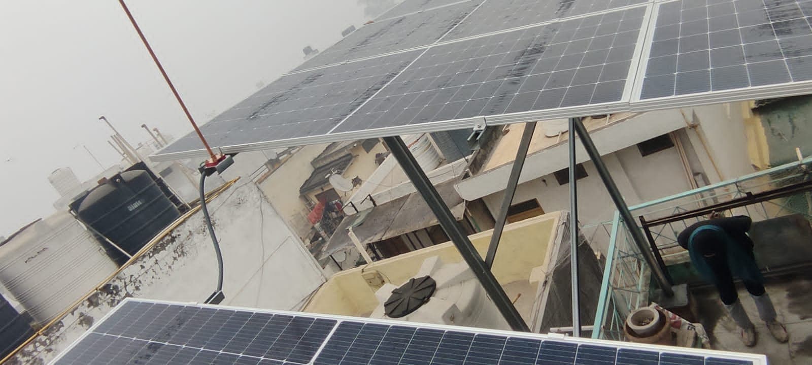 Solar panels on an elevated steel structure above a rooftop, with water tanks and neighbouring houses below