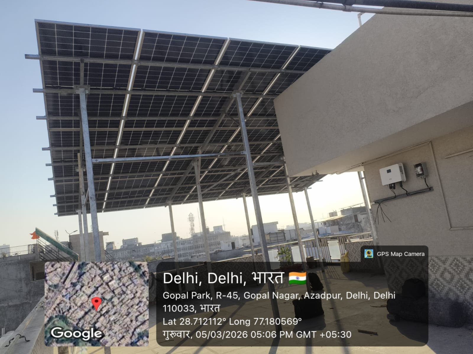 Underside of a large elevated solar array on galvanised steel posts with a wall-mounted inverter on a Delhi rooftop