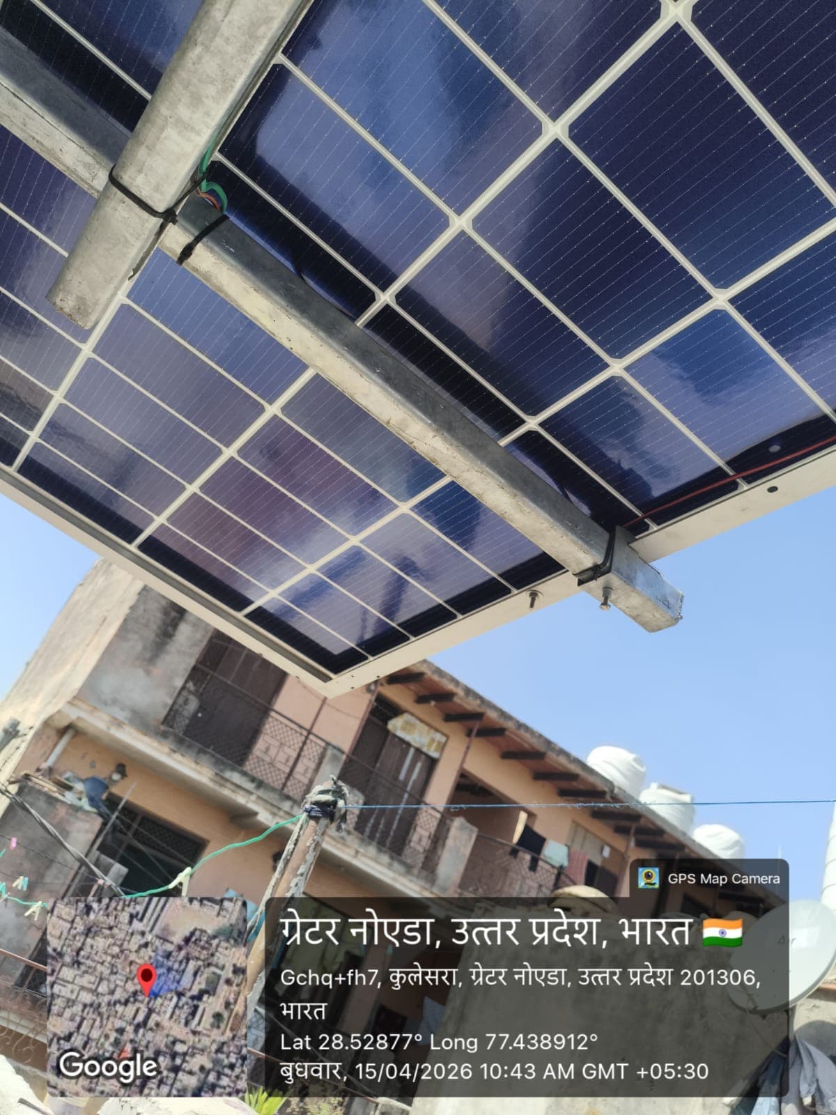 Close-up underside of solar panels clamped to galvanised rails, looking up past a neighbouring building in Greater Noida