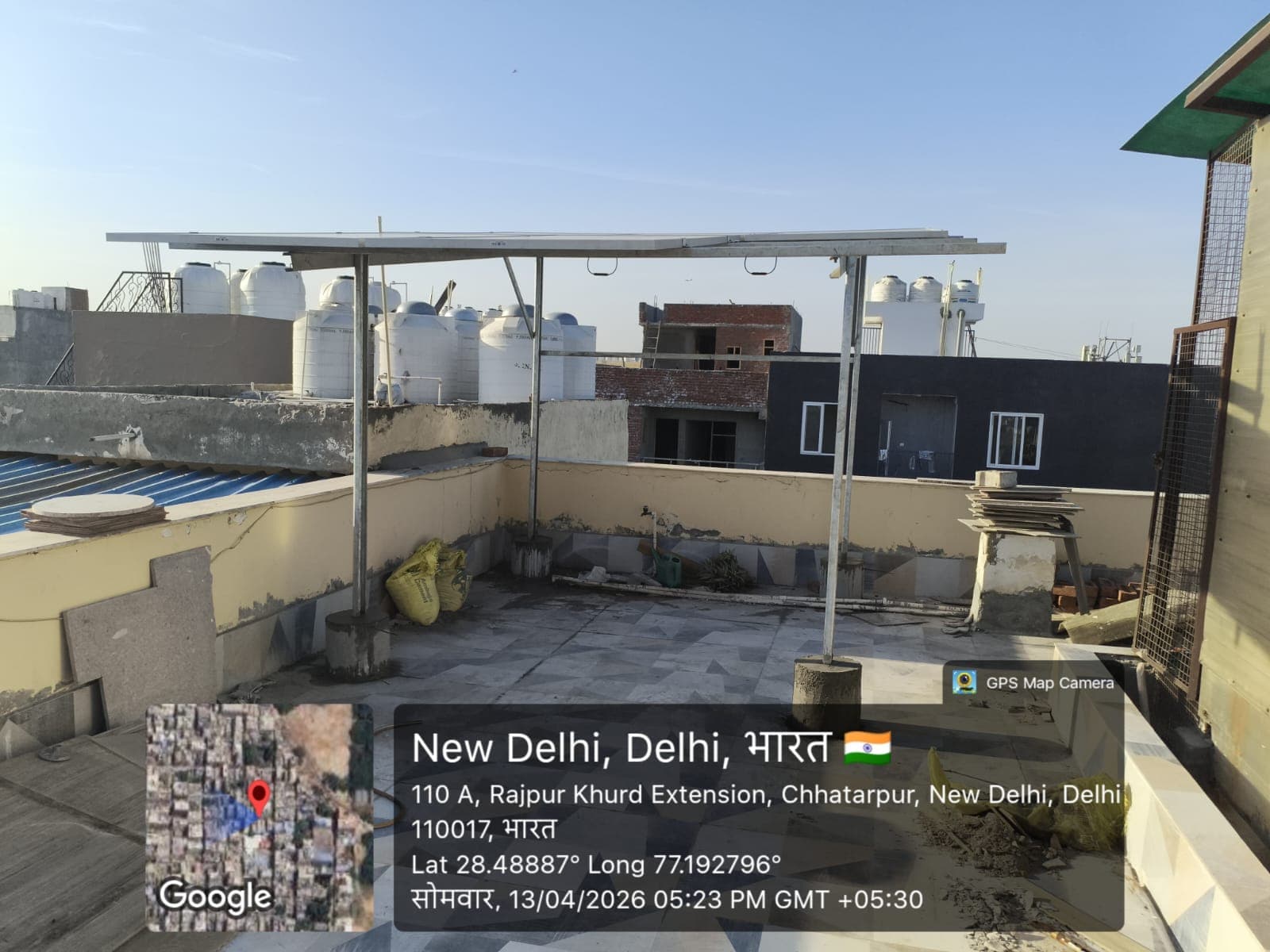 Elevated solar panel structure on steel posts above a tiled rooftop with neighbouring water tanks in New Delhi