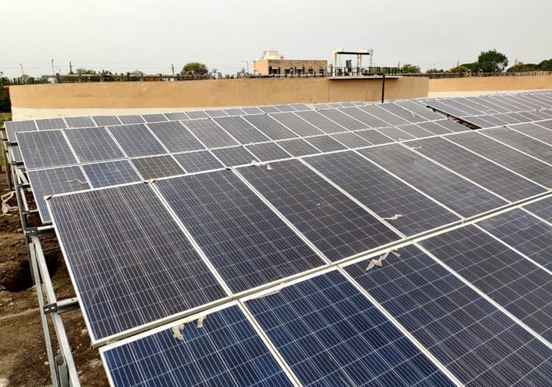 Rows of polycrystalline solar panels on low steel rails inside a walled compound, several streaked with bird droppings, under an overcast sky