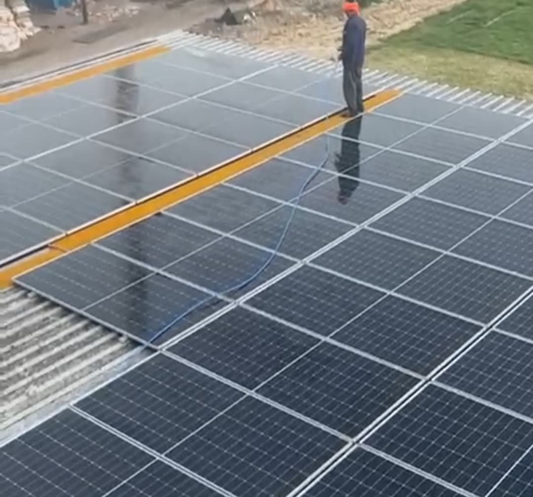 A worker in an orange cap stands on a walkway between rows of wet solar panels on a metal-sheet roof, holding a hose, with open ground and grass beyond