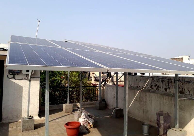 Rows of solar panels on an elevated galvanised steel structure above a flat concrete rooftop, with a wall-mounted inverter on the left wall and a parapet behind