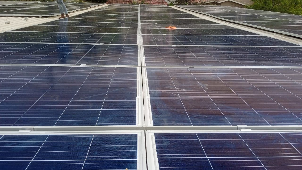 Rows of solar panels on a rooftop, with a person standing at the far end and trees behind