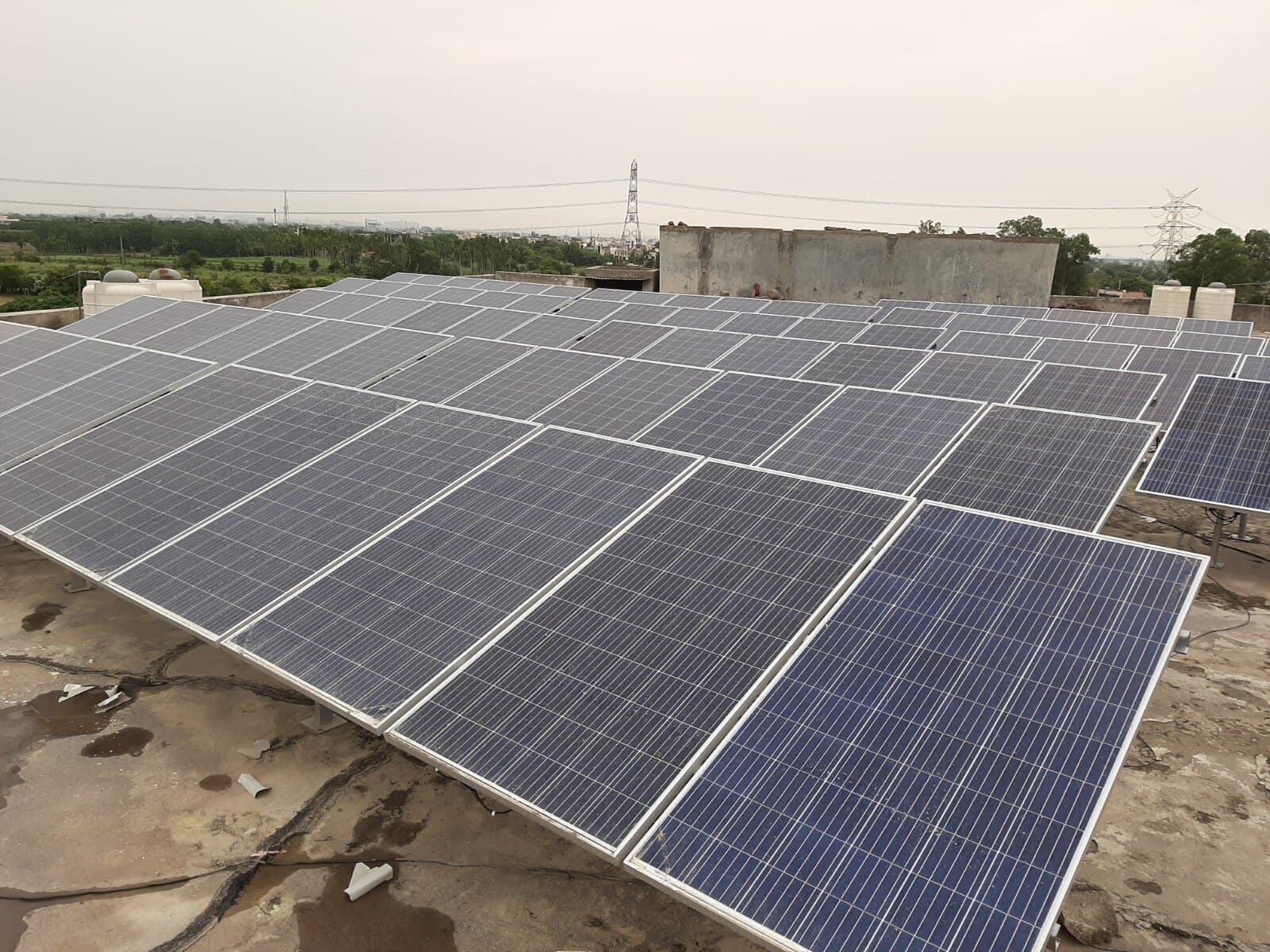Rows of solar panels on tilted frames across a flat concrete roof, with a parapet wall, water tanks, trees and two transmission towers behind them under an overcast sky.