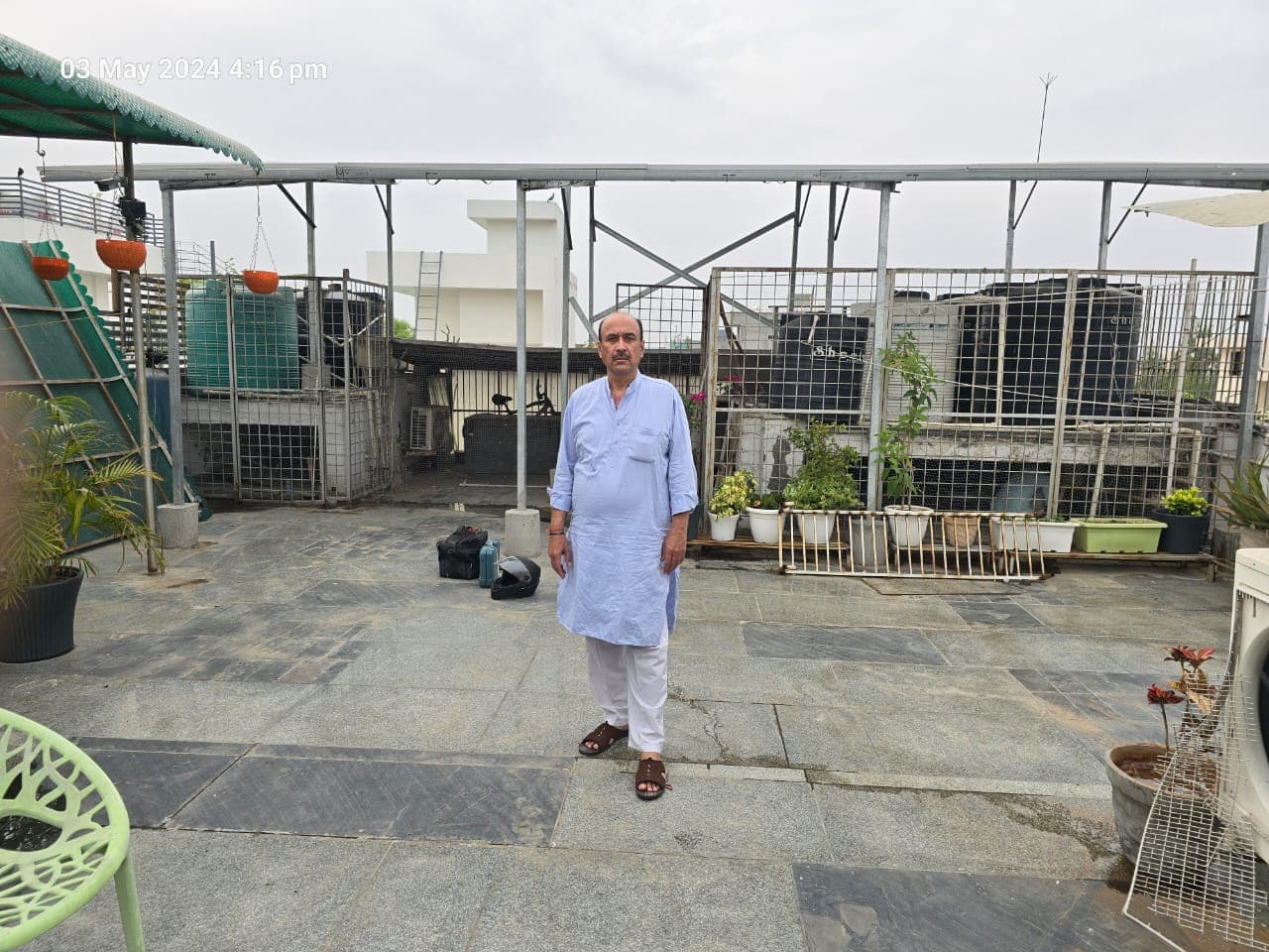 A man in a light blue kurta standing on a stone-paved rooftop terrace beneath a tall galvanised steel frame, with water tanks and air-conditioner outdoor units in wire-mesh enclosures, potted plants and a green shade canopy behind him