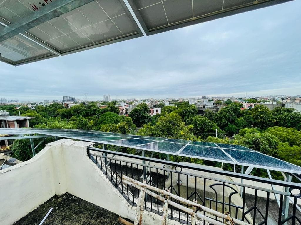 A row of solar modules on a raised steel structure above a rooftop parapet and black railing, with trees and a city skyline under an overcast sky, and more modules overhead