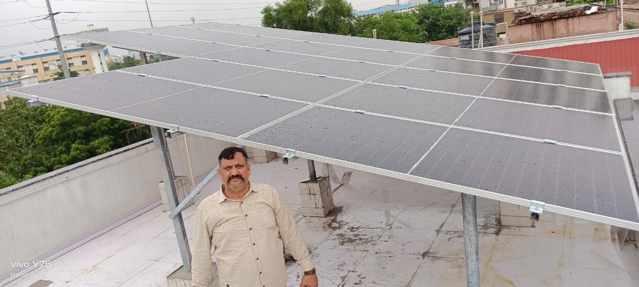 A man in a light shirt standing on a flat terrace beneath rows of solar modules mounted on a raised steel structure, with trees, buildings and a red tin roof behind