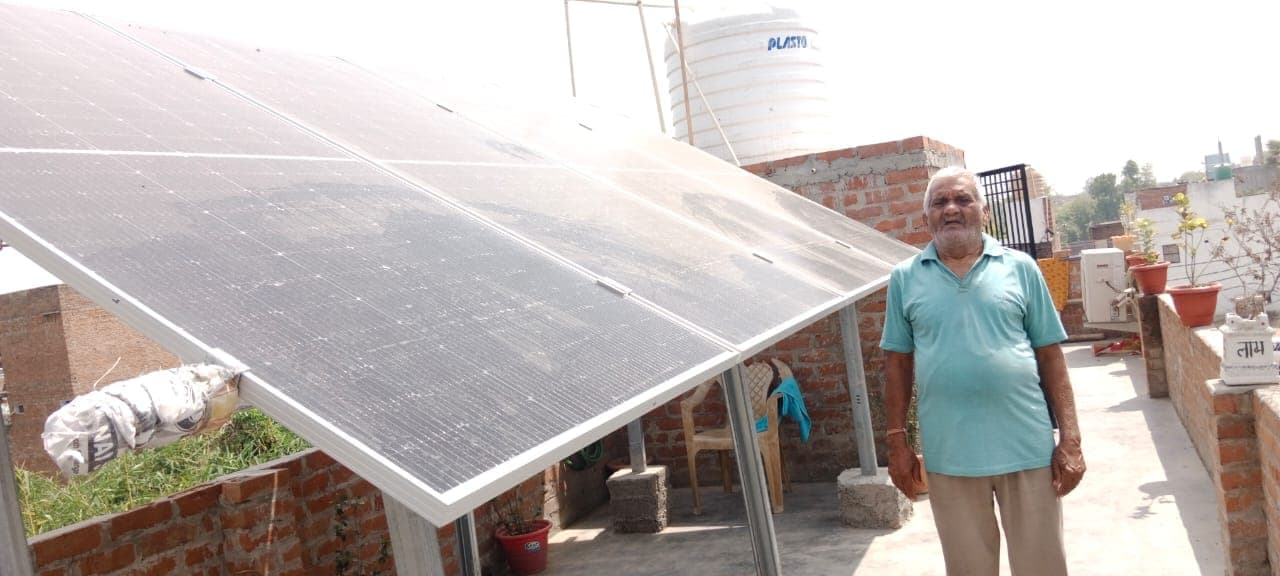 Solar panels on a raised metal frame on a brick-walled rooftop, with a white water tank behind them and a man standing beside the structure.