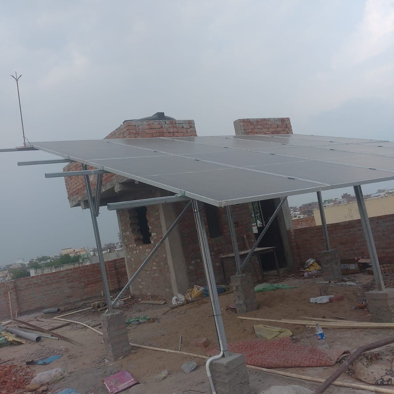 Solar panels on a raised steel structure above a rooftop with brick parapet walls, a water tank and the city behind