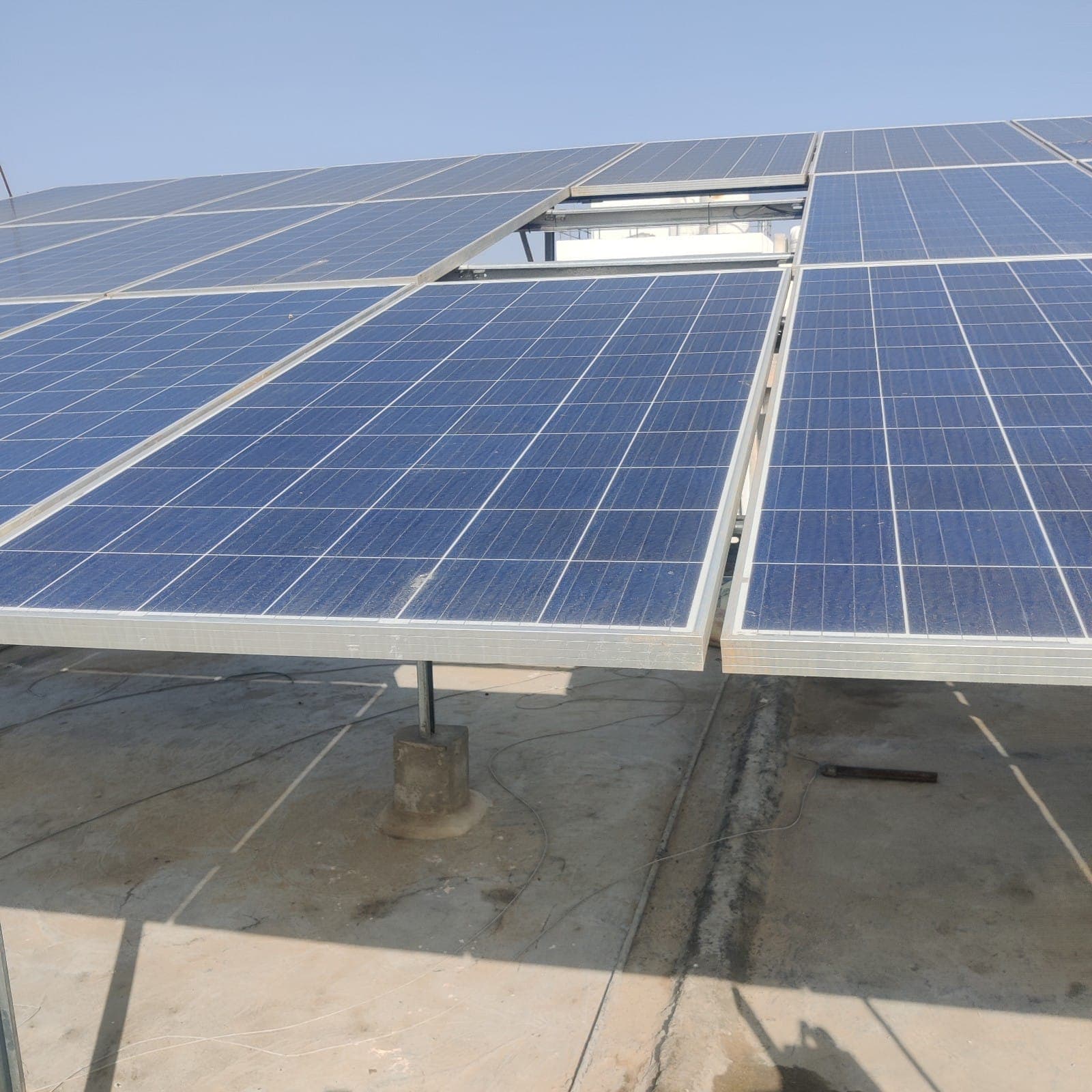 Rows of solar panels on an elevated galvanised steel structure above a concrete terrace, with a white rooftop tank behind.